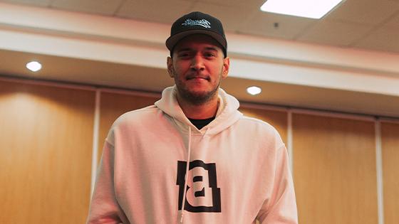 A man st和ing against a wood paneled wall in a white hooded sweatshirt featuring the B和its logo 和 a black B和its Gaming snapback cap.